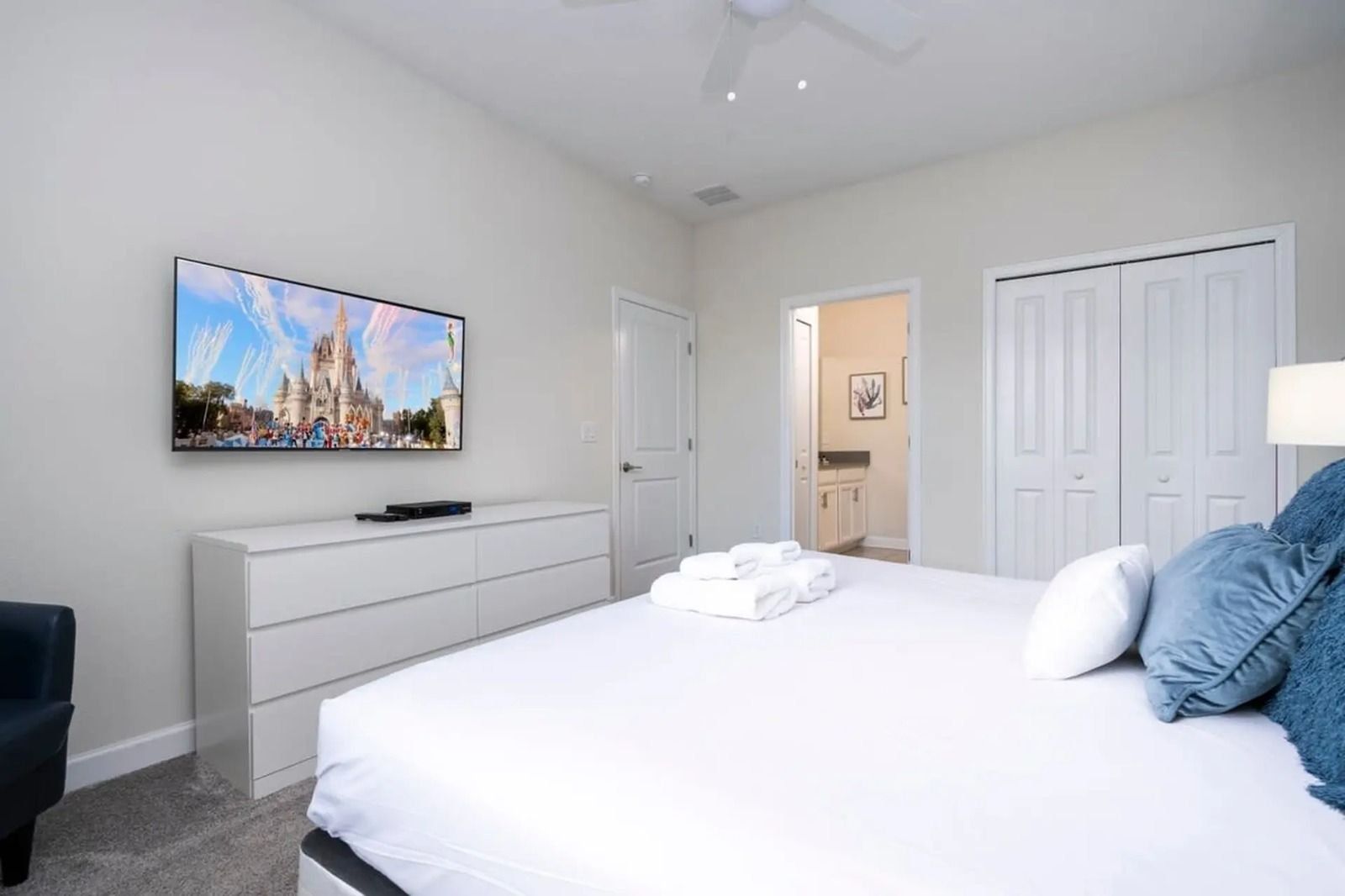 Une chambre avec un grand lit, un téléviseur à écran plat fixé au mur, une commode et une porte donnant sur une autre pièce.