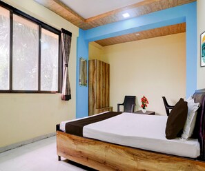 A bedroom with a wooden bed frame, a window with curtains, a chair, a small table with a vase of flowers, and a wardrobe. - Classic Hotel Shivneri (Vasai)