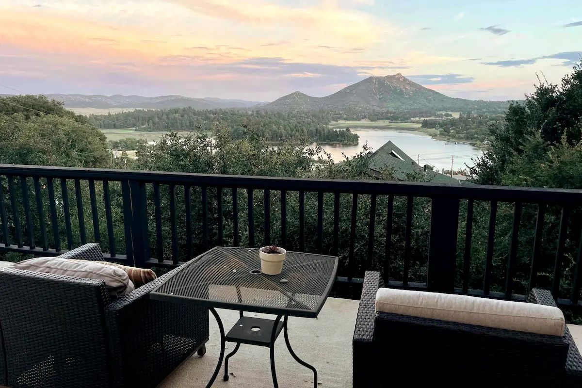 A balcony with wicker furniture, a table with a potted plant, and a scenic view of a lake and mountains.