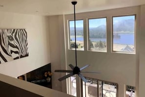 Ein Zimmer mit Deckenventilator, großen Fenstern mit Blick auf einen See und Berge sowie einem Zebrastreifen-Kunstwerk an der Wand.