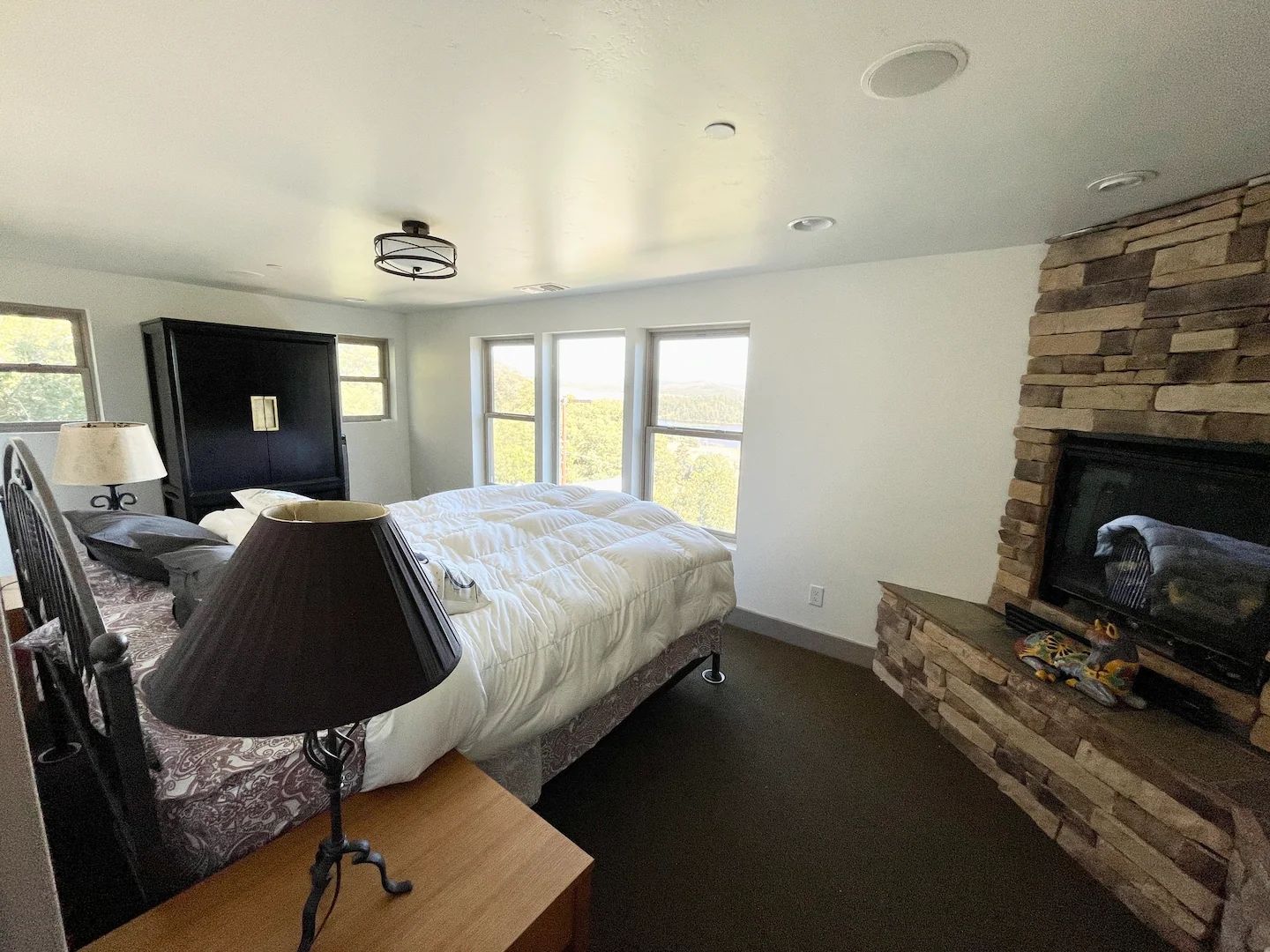 A bedroom with a bed, a nightstand, a lamp, a dresser, a fireplace, and a large window.