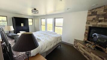 A bedroom with a bed, a nightstand, a lamp, a dresser, a fireplace, and a large window.