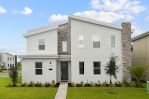 A modern two-story house with a stone facade, a front entrance, and a small front yard with young trees. - 9 Bedroom Villa With Pool at Storey Lake (Kissimmee)