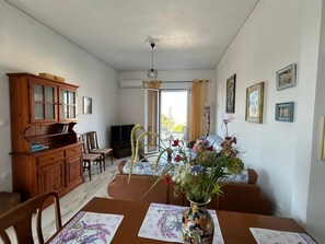 A dining area with a wooden cabinet, chairs, a table with a vase of flowers, and a television in the background. - Family Apartment 150 meters from the beach (Selinia)
