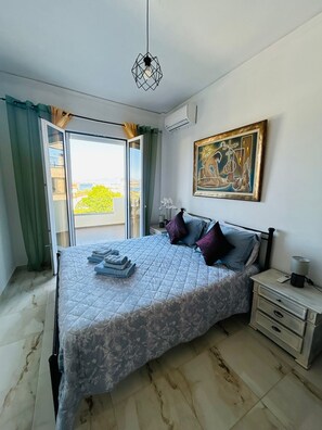 A bedroom with a bed, a nightstand, a painting, and a view through a window. - Family Apartment 150 meters from the beach (Selinia)