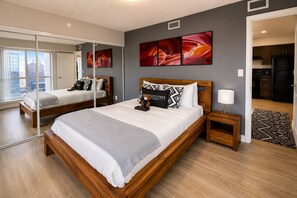 Ein modernes Schlafzimmer mit einem großen Bett, einem Nachttisch, einer Stehlampe und Blick auf die Stadt durch große Fenster.