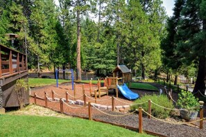 A playground with a slide, climbing structures, and a small wooden playhouse surrounded by trees. - Sleeps 12 | 3 Baths | Hot Tub | Near Bear Valley (Arnold)