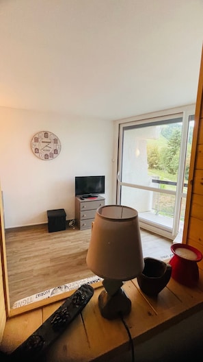 Ein Zimmer mit Fernseher, einer Uhr, einer Lampe und einem Fenster mit Blick auf Grünflächen.