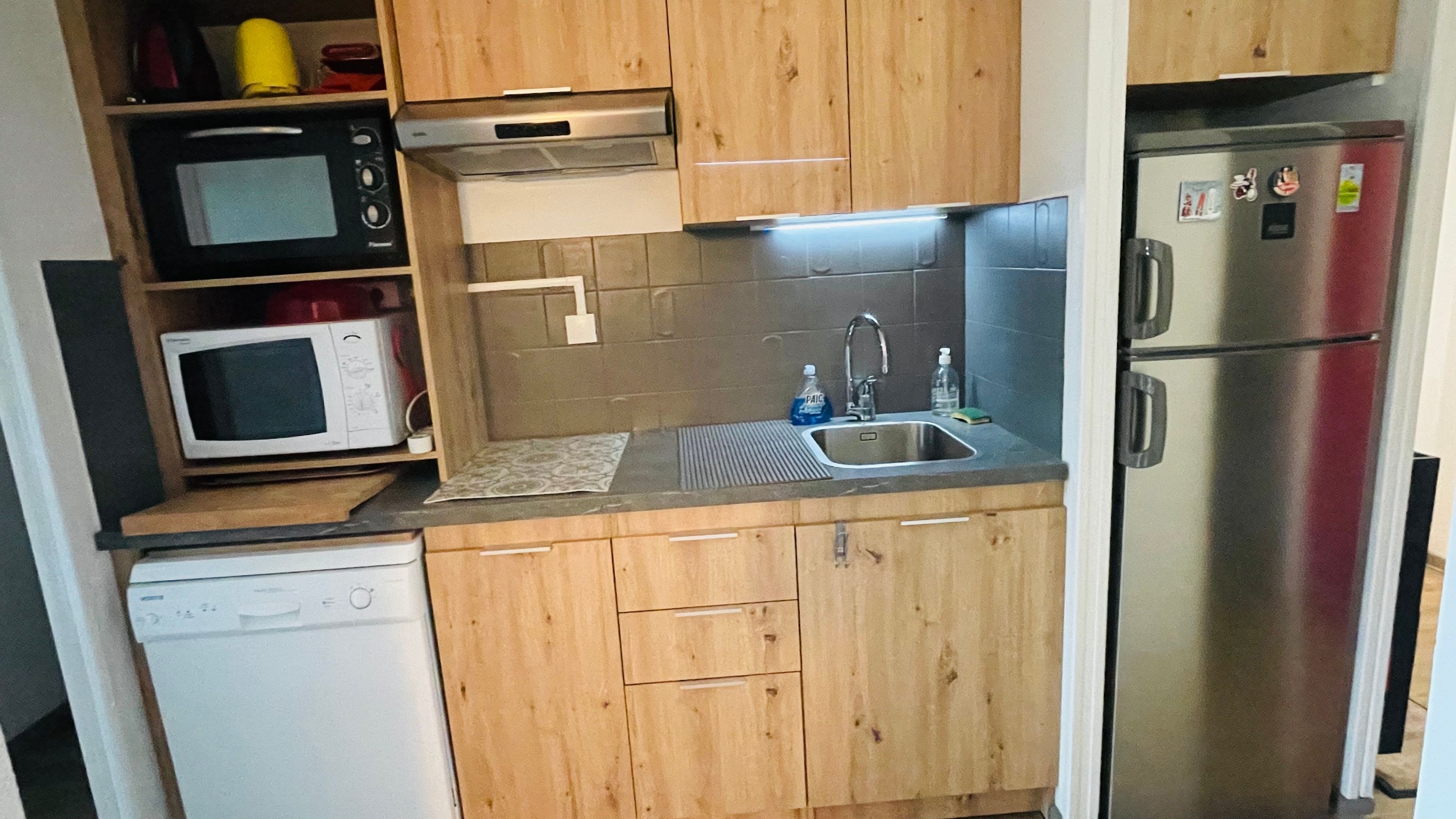 Eine kleine Küche mit Holzausstattung, einem Edelstahlkühlschrank, einer Mikrowelle und einem Geschirrspüler.