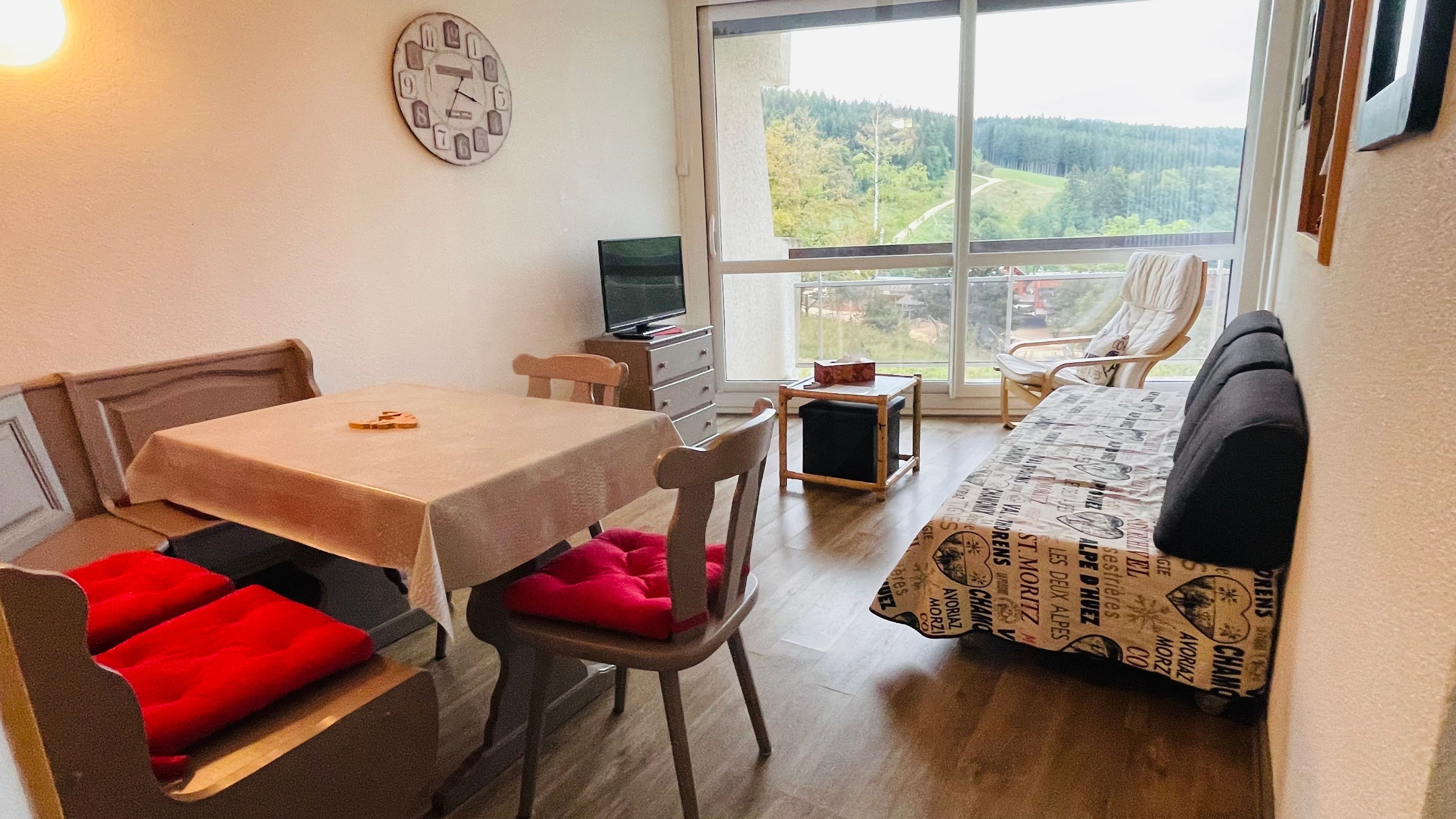 Ein gemütliches Zimmer mit Esstisch, Stühlen, einer Couch, einem Fernseher und einem großen Fenster mit Blick ins Freie.