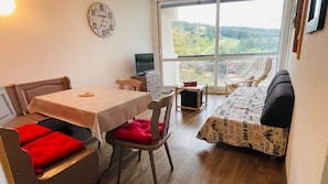 Ein gemütliches Zimmer mit Esstisch, Stühlen, einer Couch, einem Fernseher und einem großen Fenster mit Blick ins Freie.