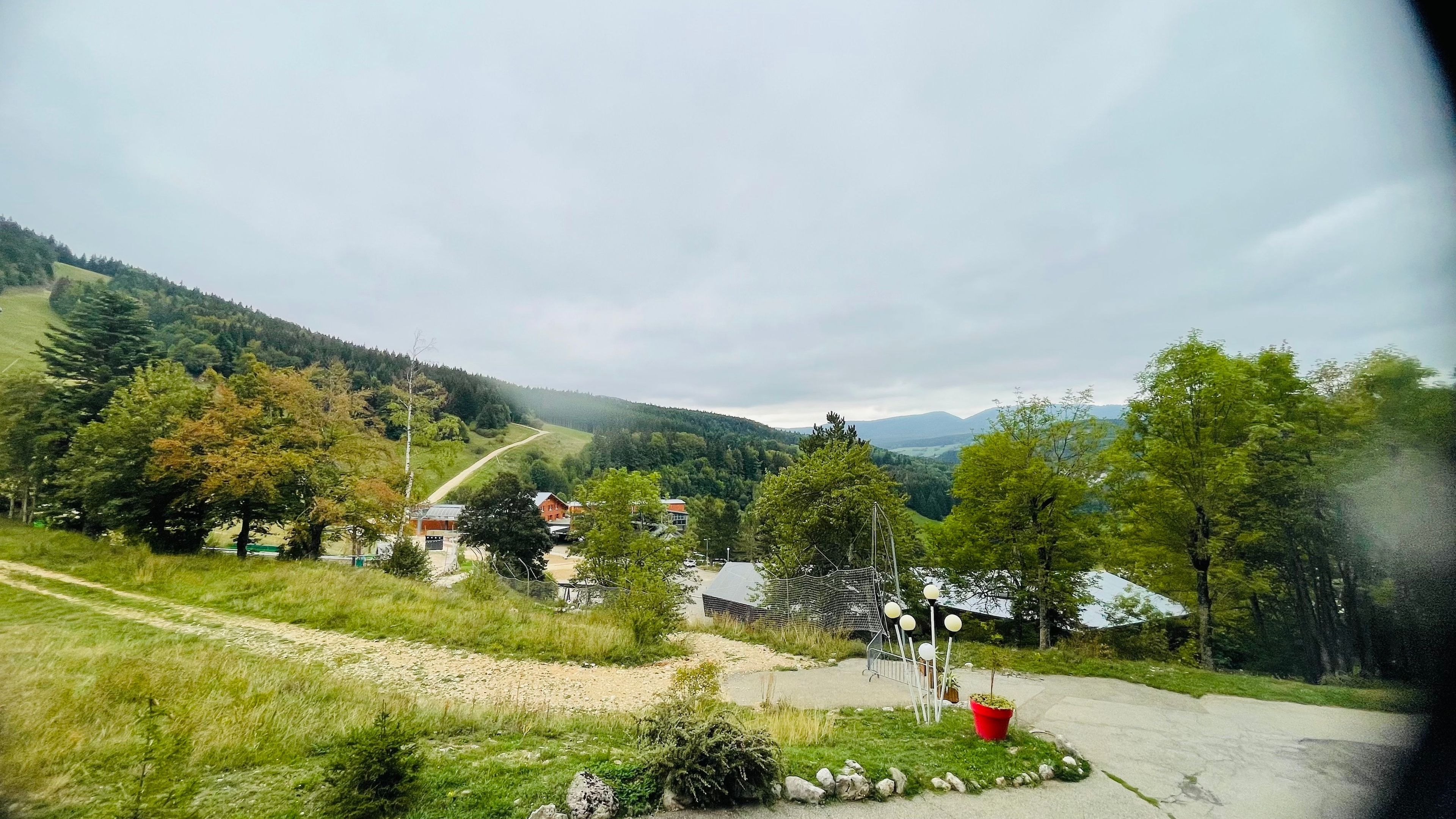 Ländliche Landschaft mit Feldweg, einem kleinen Gebäude und einem bewaldeten Hügel im Hintergrund.