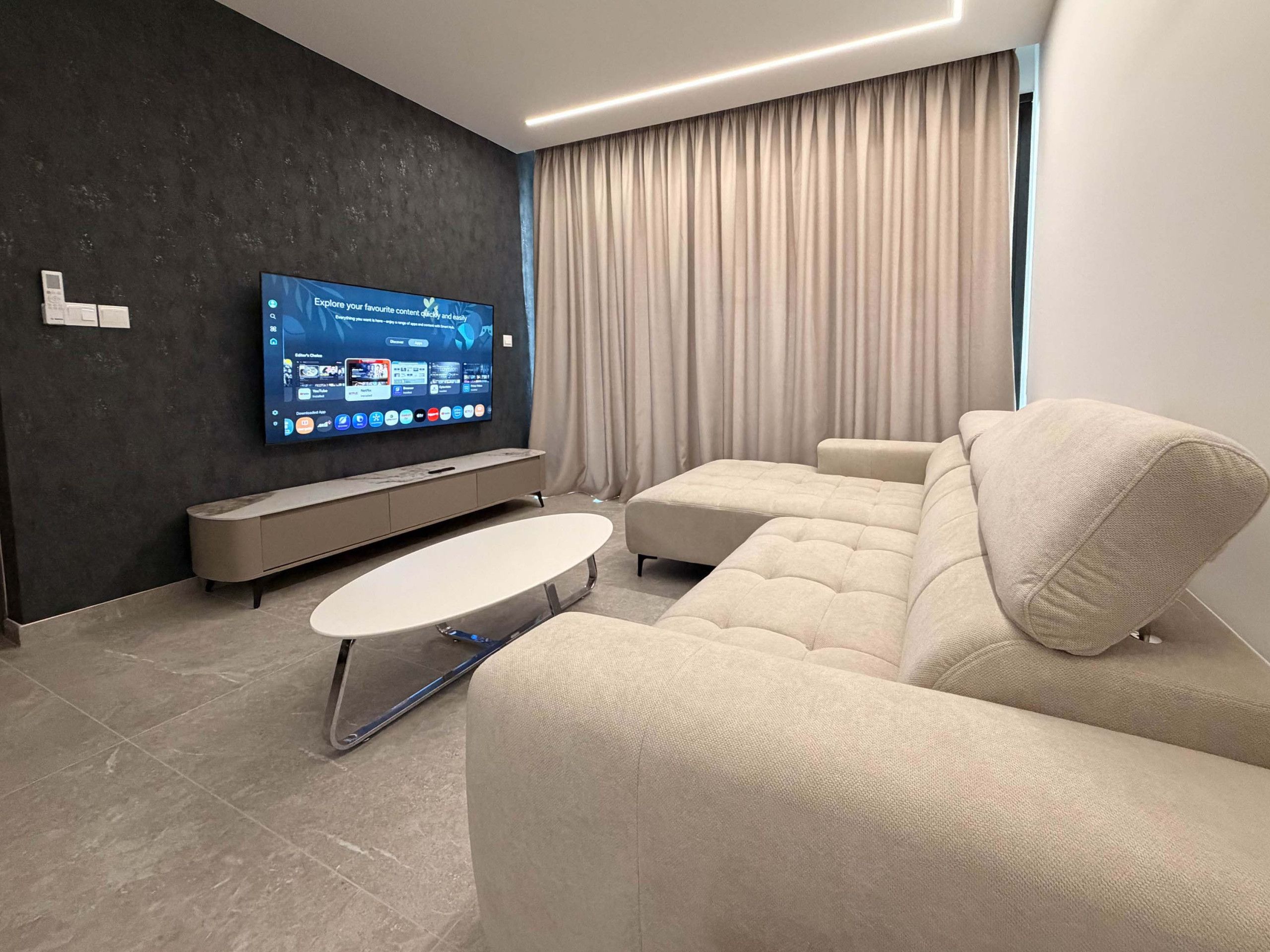 A modern living room with a flat-screen TV mounted on a dark wall, a beige sofa, a coffee table, and a large window with curtains.