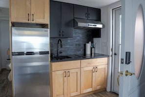 A modern kitchen with a stainless steel refrigerator, black cabinets, a black countertop, and a stainless steel range hood. - Hamptons Oceanfront Oasis (Hampton Bays)