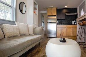 A modern living room with a sofa, a round coffee table, and a kitchen area with wooden cabinets and a stainless steel refrigerator. - Hamptons Oceanfront Oasis (Hampton Bays)