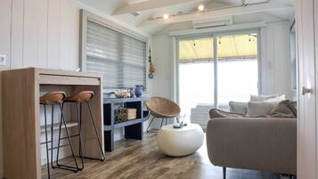 A modern living space with a kitchen area, a dining table with stools, a sofa, and a large window providing natural light.