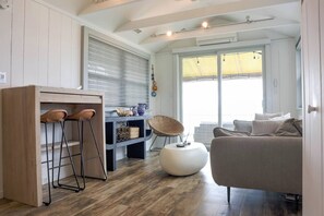 A modern living space with a kitchen area, a dining table with stools, a sofa, and a large window providing natural light. - Hamptons Oceanfront Oasis (Hampton Bays)