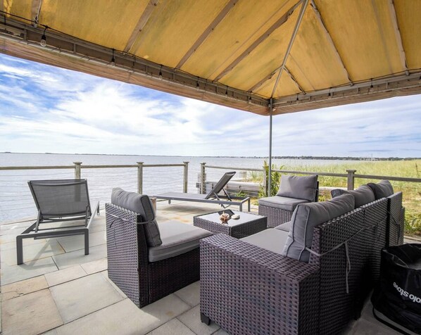 An outdoor patio with wicker furniture, a table, and a view of the water. - Hamptons Oceanfront Oasis (Hampton Bays)