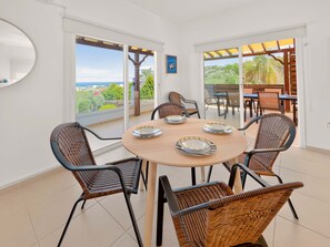 Un coin repas avec une table ronde et six chaises en osier, de grandes portes vitrées donnant sur un patio extérieur avec vue sur le paysage.