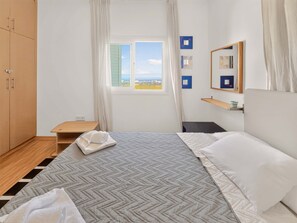 A hotel room with a bed, a window with a view, a wooden wardrobe, and a small table. - Saint Elias Skyview | Hilltop Villa & Garden (Protaras)
