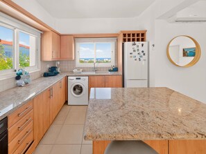 A compact kitchen with a washing machine, a refrigerator, and a window with a view of buildings. - Saint Elias Skyview | Hilltop Villa & Garden (Protaras)