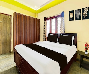 A bedroom with a bed, a wooden wardrobe, a window with curtains, and a nightstand with a vase of flowers. - Classic Akshaya Kannur (Penumuluru)