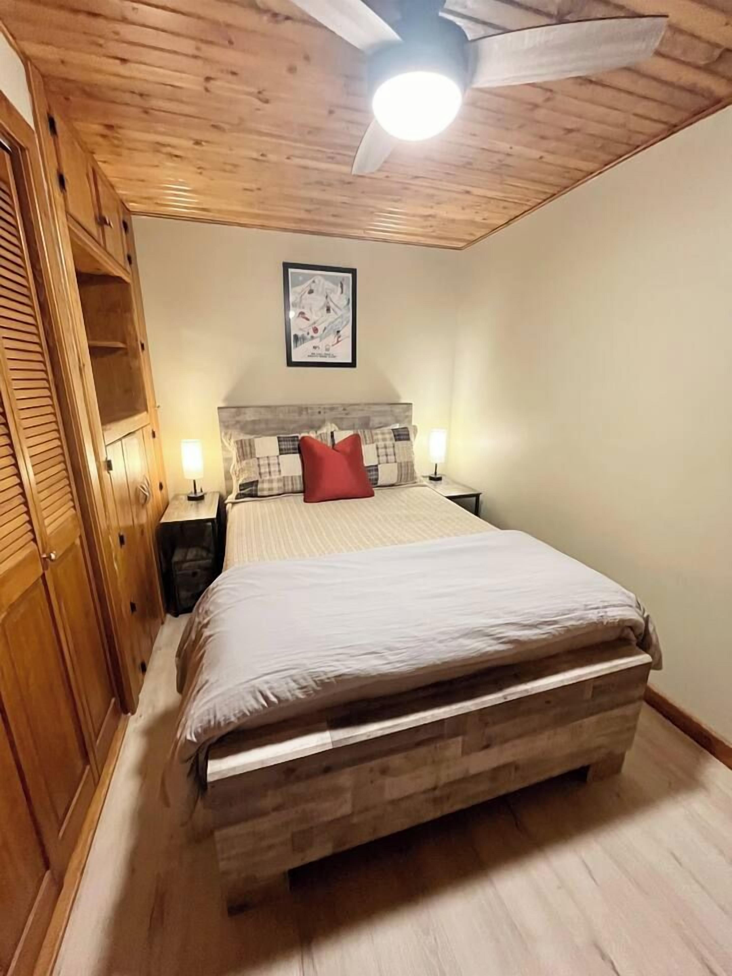 Une chambre à coucher avec un plafond en bois, un lit doté d’une tête de lit, deux tables de chevet et un luminaire fixé au mur.