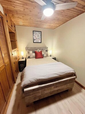 Une chambre à coucher avec un plafond en bois, un lit doté d’une tête de lit, deux tables de chevet et un luminaire fixé au mur.