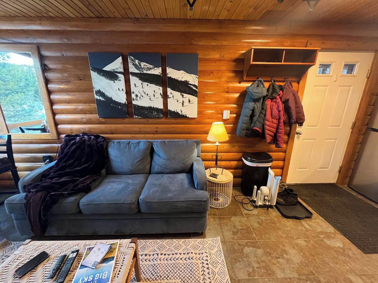 Het interieur van een blokhut met een bank, een lamp, een bijzettafeltje en een deur met jassen er aan.
