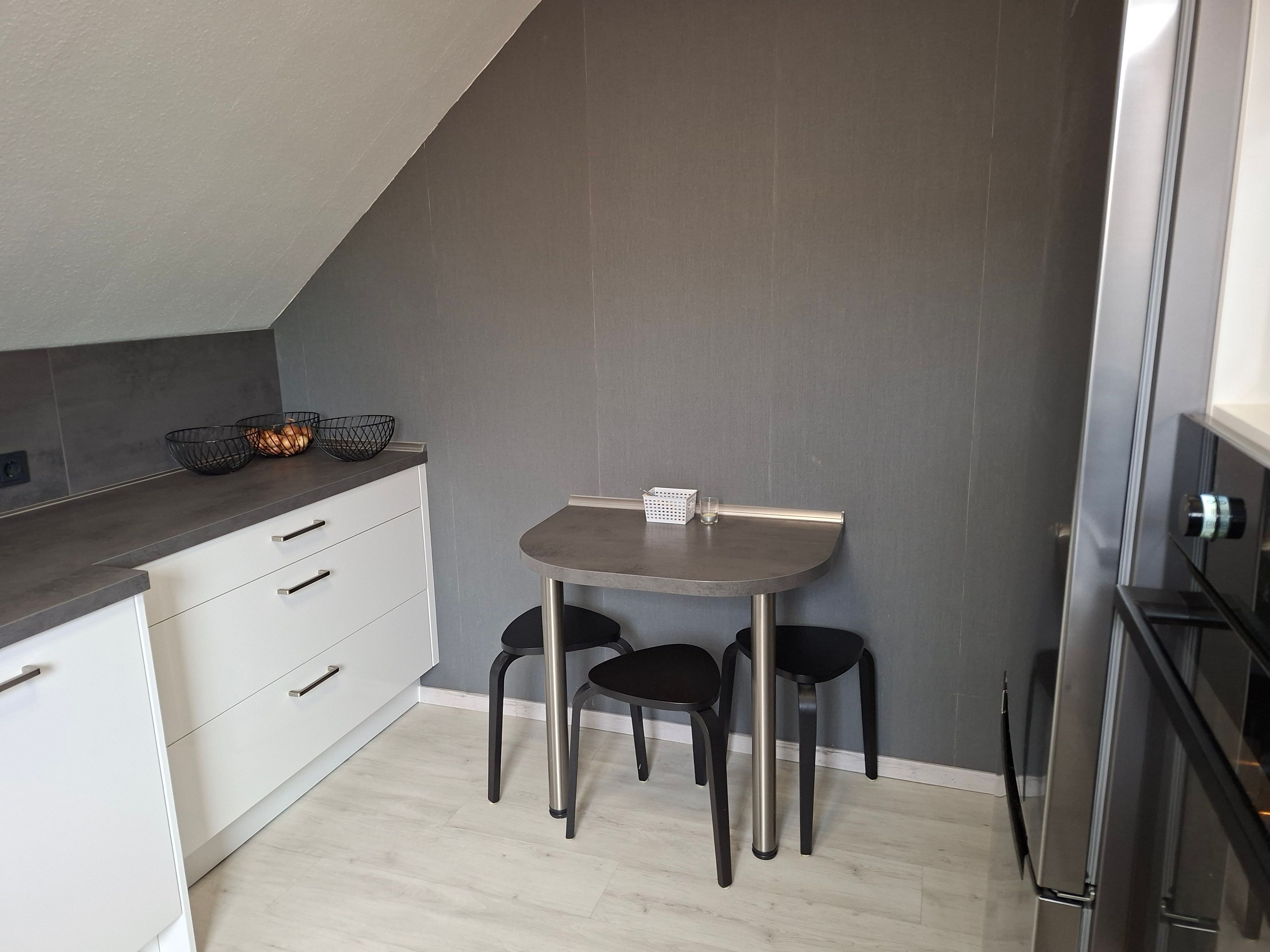 Cocina compacta con una pequeña mesa de comedor y dos sillas, un armario blanco con cajones y un refrigerador moderno.