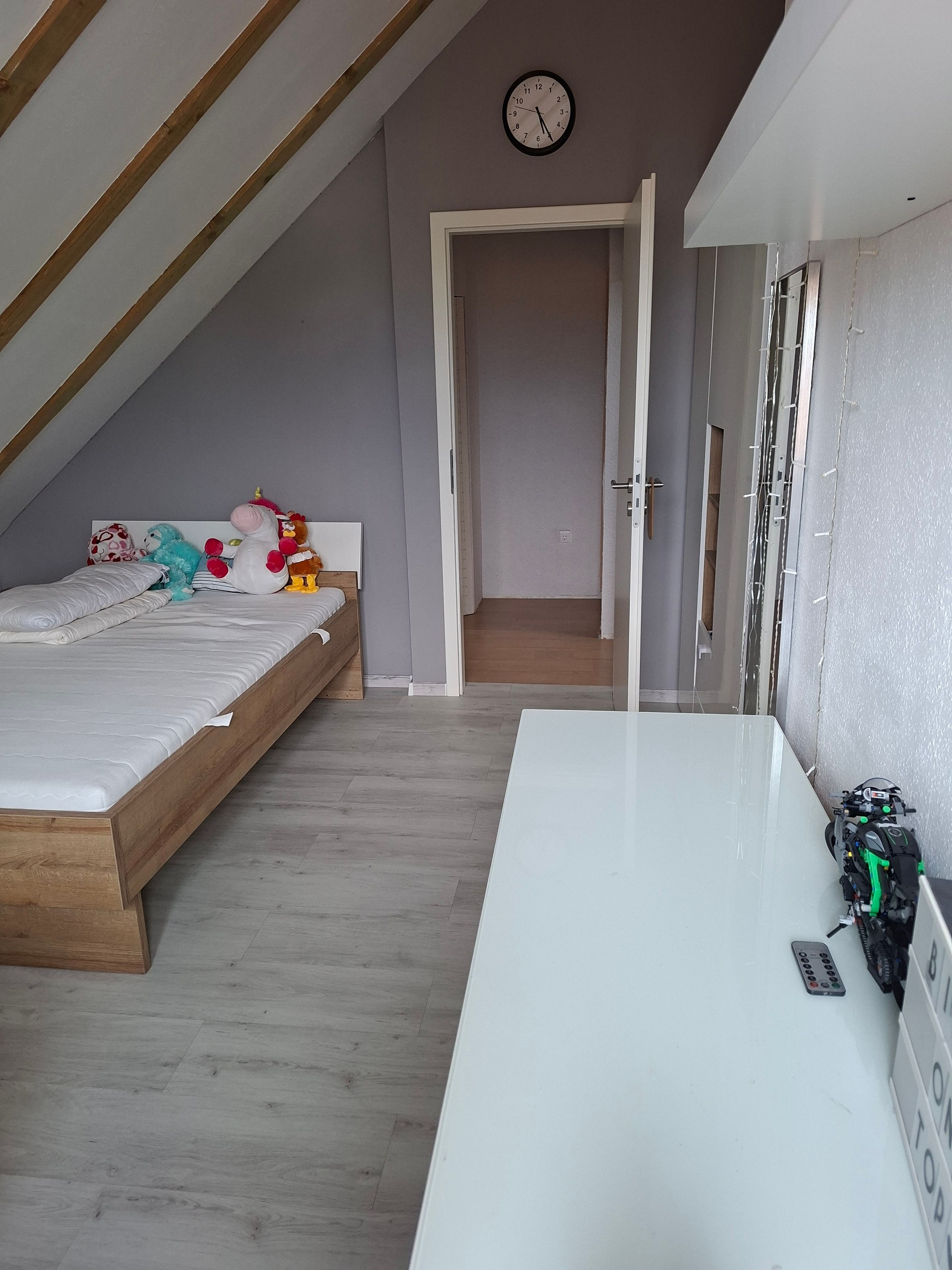 Ein Schlafzimmer mit schrägem Dach, einem Bett mit Kuscheltieren, einem weißen Schreibtisch mit Rucksack und einer Uhr an der Wand.