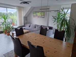 Ein modernes Wohnzimmer mit einem grauen Sofa, einem Holzesstisch und schwarzen Stühlen. Übertöpfe und ein Gemälde sind an der Wand zu sehen.