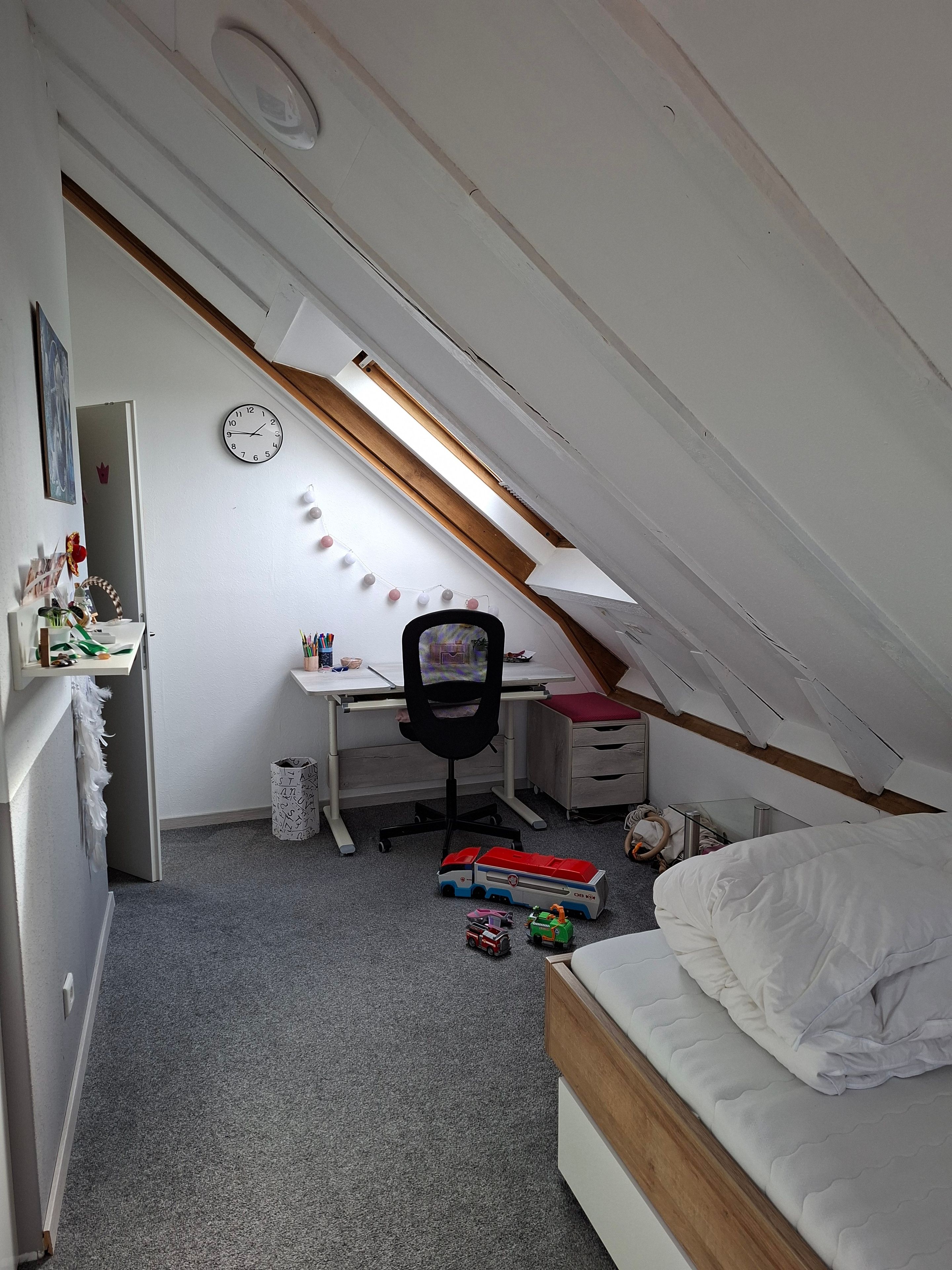 Ein Zimmer mit schrägen Wänden, einem Schreibtisch mit Stuhl, einem Bett und Spielzeug auf dem Boden.