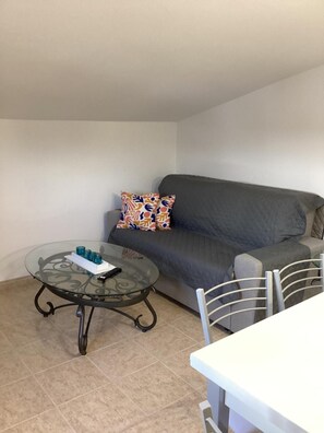 A living room with a glass coffee table, a grey sofa, colorful pillows, and a white dining table. - Apartment 'Vista Mar' with Sea View, Wi-Fi and Air Conditioning (Lourinhã)