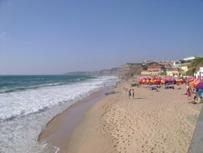 Ein Strand mit Sonnenschirmen und Personen, ein klarer Himmel und Gebäude im Hintergrund.