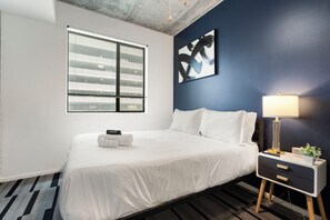 Ein modernes Hotelzimmer mit einem großen Bett, einem Nachttisch mit Lampe und einem Fenster mit Jalousien.
