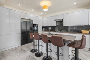 A modern kitchen with white cabinets, a stainless steel refrigerator, a built-in oven, and a bar area with stools. - Stylish 4BR Executive Home/Huge Kitchen/Sleeps7 (Bolton)