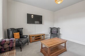 Un salon avec un téléviseur à écran plat fixé au mur, deux fauteuils en cuir, une table basse en bois et une petite table d’appoint en bois avec des paniers.