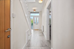 Un couloir avec une porte en bois, des murs blancs et un sol carrelé, qui mène à une porte dotée d’un panneau de verre.