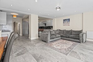 A modern living room with a grey sectional sofa, a dining table with chairs, and a kitchen in the background. - Stylish 4BR Executive Home/Huge Kitchen/Sleeps7 (Bolton)