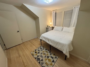A bedroom with a bed, a bedside table, a lamp, a window with blinds, a door, and a patterned rug. - Top Tier Private Duplex, Centrally Located! (Superior)