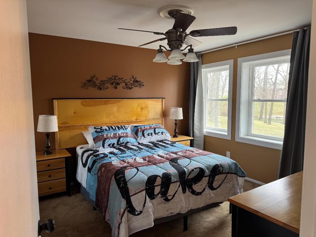 A bedroom with a bed, a ceiling fan, a nightstand, a lamp, and a window with curtains.