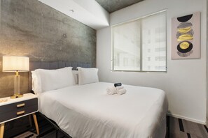A hotel room with a bed, a bedside table with a lamp, a window with blinds, and a wall with a textured design. - Classy 2BR | Pool + W/D | Near Arts District (Phoenix)
