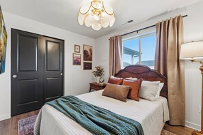 A bedroom with a large bed, a wooden headboard, a window with curtains, a chandelier, and a door. - Boutique Style. Cozy 2BR+ Office for Two. Fenced yard. 30+ days stays. Near I-84 (Nampa)