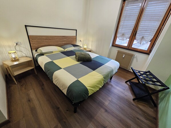 Een slaapkamer met een bed, nachtkastjes, een lamp, een raam met zonwering en een stoel.