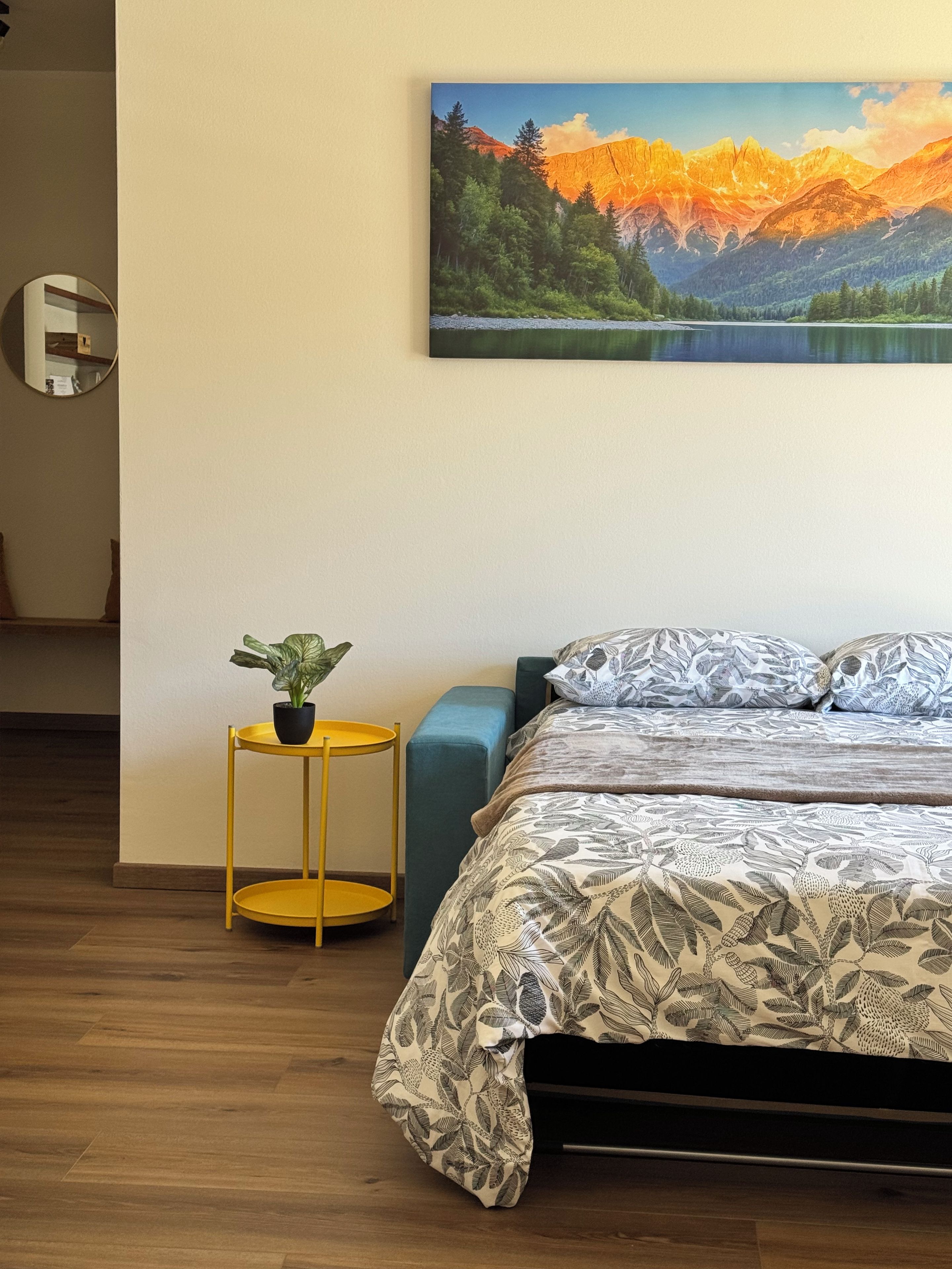A bedroom with a bed, a small round yellow table with a plant, and a large landscape painting on the wall.