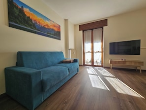 Una sala de estar con un sofá azul, una mesa de centro de madera, un televisor de pantalla plana y un cuadro de un paisaje montañoso.