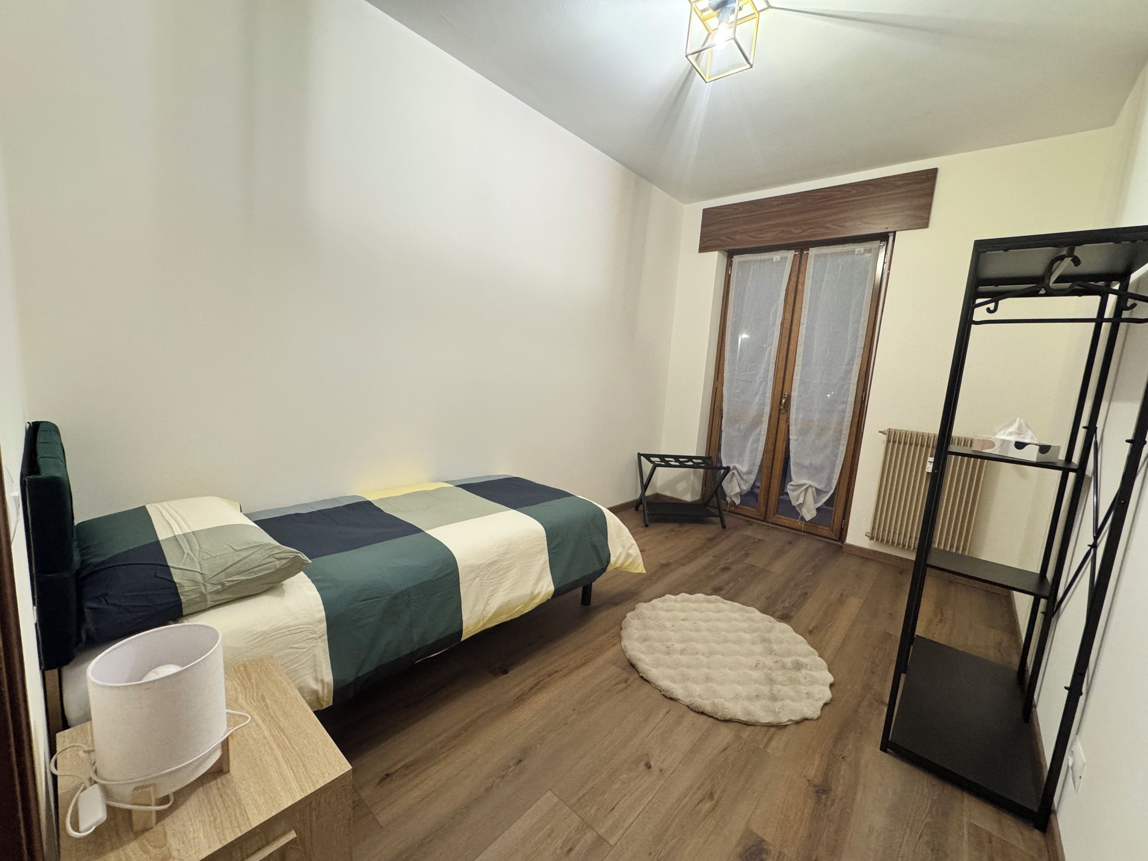 A bedroom with a bed, a bedside table, a lamp, a radiator, and a window with curtains.