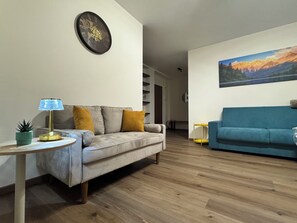 Een woonkamer met een grijze bank, een blauwe bank, een ronde wandklok en een schilderij van een berglandschap.
