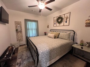 A bedroom with a bed, a ceiling fan, a window with curtains, a wall-mounted TV, and a bedside table with a lamp.